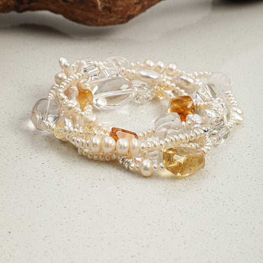 A stack of beaded stretch bracelets featuring clear faceted beads, white pearl-like beads, and amber-colored stone accents, displayed on a light speckled surface with a piece of driftwood in the background.