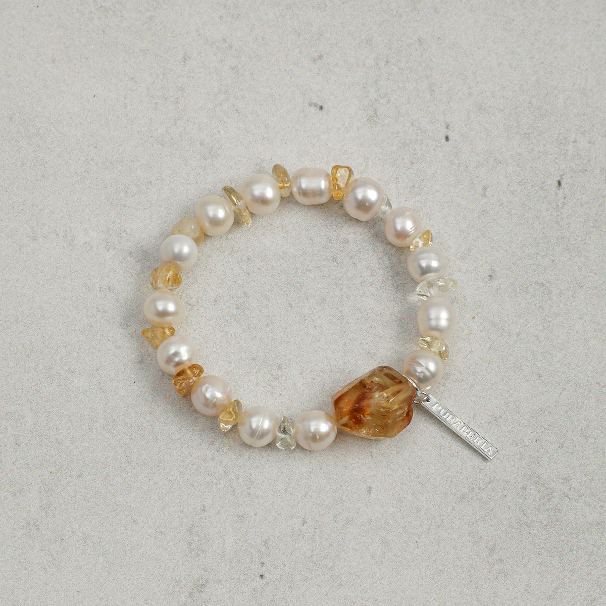 A beaded stretch bracelet featuring white freshwater pearls, small clear faceted beads, amber-orange chips, and a polished amber-orange stone centerpiece with a silver-tone tag, displayed on a textured light gray surface.