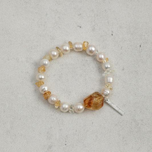 A beaded stretch bracelet featuring white freshwater pearls, small clear faceted beads, amber-orange chips, and a polished amber-orange stone centerpiece with a silver-tone tag, displayed on a textured light gray surface.