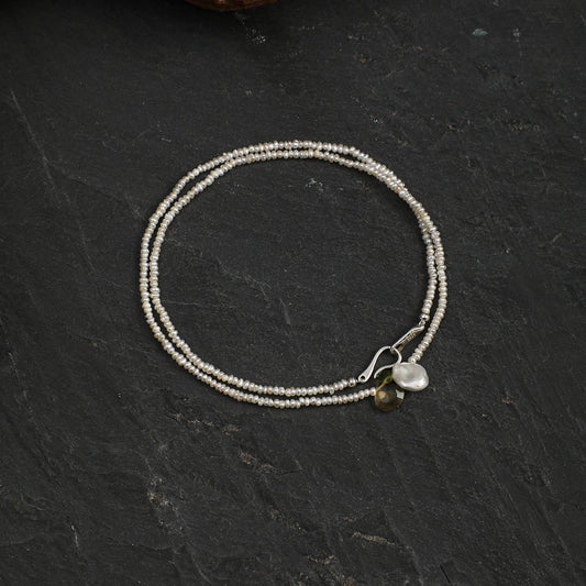 A delicate double-strand beaded bracelet in silver with a small white pearl and green bead accent, displayed on a dark slate surface.