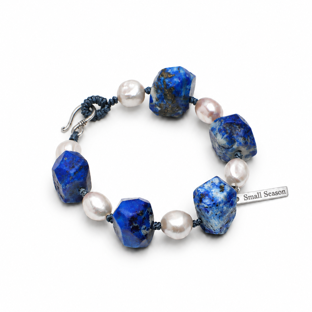 A beaded bracelet featuring textured blue lapis lazuli stones and white freshwater pearls, accented by a dark blue cord and a silver toggle clasp. A small silver tag on the right reads 'Small Season'.