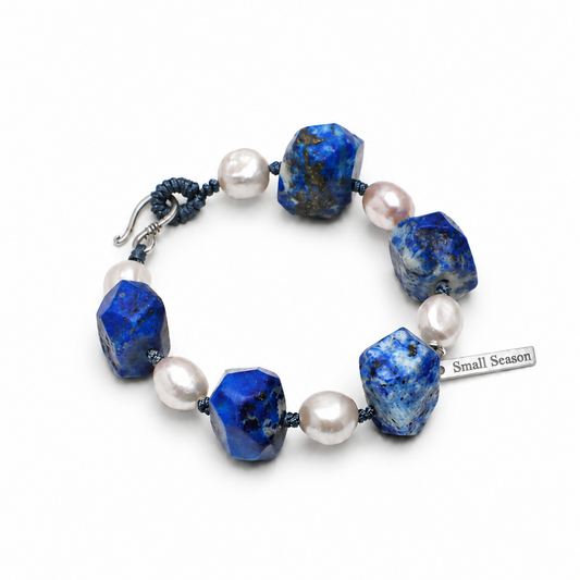 A beaded bracelet featuring textured blue lapis lazuli stones and white freshwater pearls, accented by a dark blue cord and a silver toggle clasp. A small silver tag on the right reads 'Small Season'.