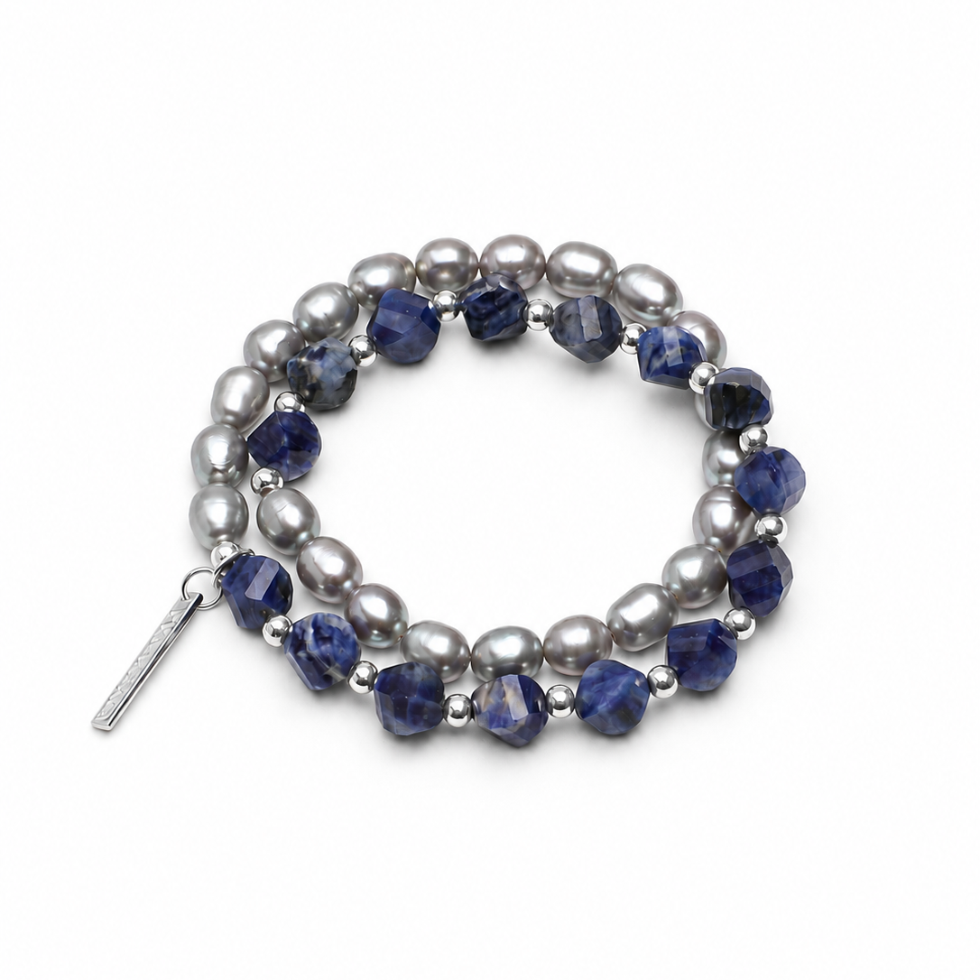 A double-strand beaded stretch bracelet featuring dark blue faceted beads and silver-gray baroque pearls, accented with small silver spacer beads and a silver toggle clasp, shown on a light cream background.