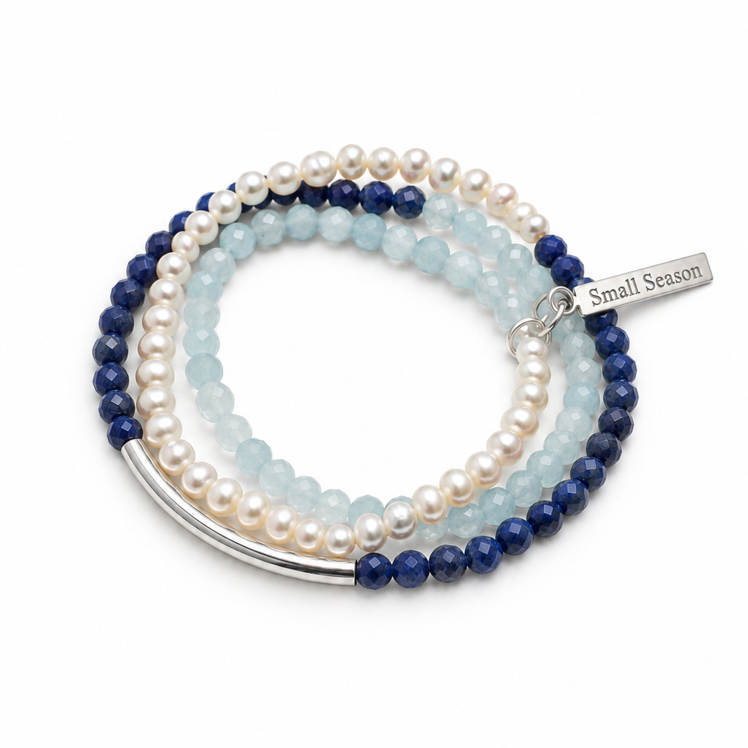 A stack of three beaded stretch bracelets on a white background. The bracelets feature white freshwater pearls, deep navy blue beads, and light blue aventurine beads, with a silver-tone curved bar and a small silver tag engraved with 'Small Season'.